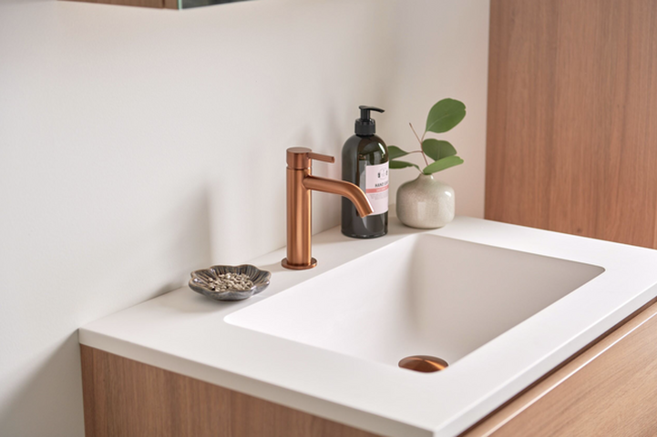 Moderne badkamer met witte wastafel en gekleurde kraan in geborsteld brons, gecombineerd met houten meubel en accessoires.