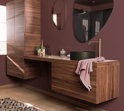 Badkamer trend met notenhouten badkamermeubel, zwarte opzetkom, koperen kraan en organische spiegels tegen bordeaux muur.