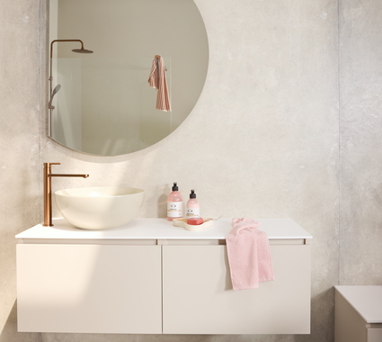 Badkamermeubel met halfronde spiegel in een rustige badkamer. Beige meubel met beige opzet waskom en bronskleurige wastafelkraan. In de reflectie van de spiegel zie je de douche.
