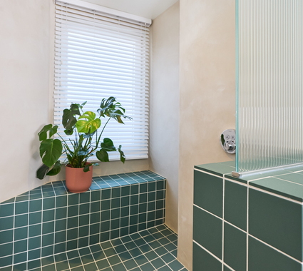 Douche met groene tegels en wit voegsel, ingebouwde zitbank, regendouche en plant bij raam met jaloezieën.