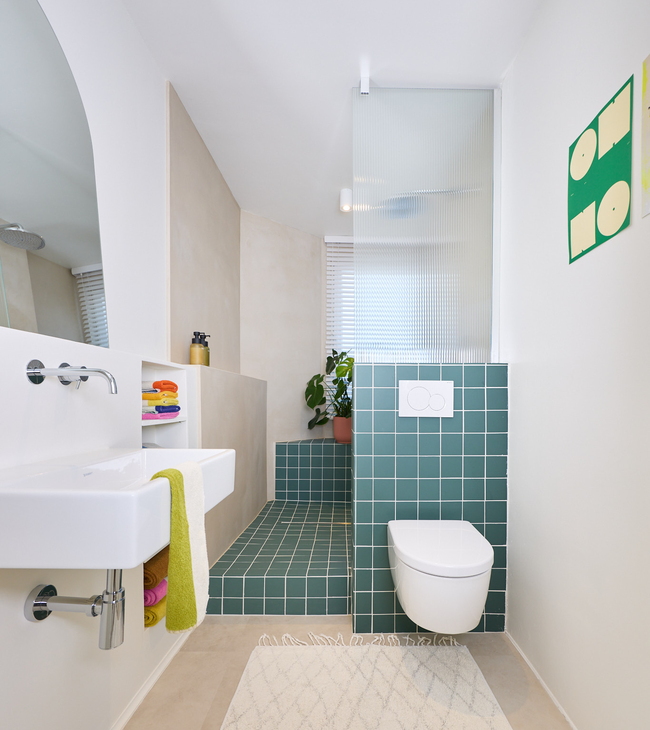 Moderne groene badkamer met wandtoilet, open douche met groene tegels en witte wastafel met kleurrijke handdoeken.