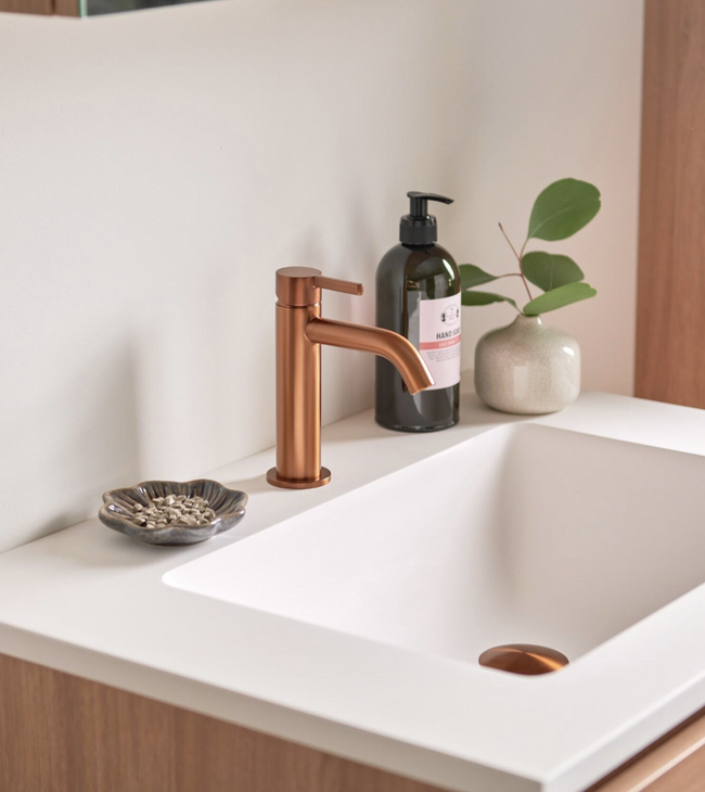 Moderne badkamer met witte wastafel en gekleurde kraan in geborsteld brons, gecombineerd met houten meubel en accessoires.