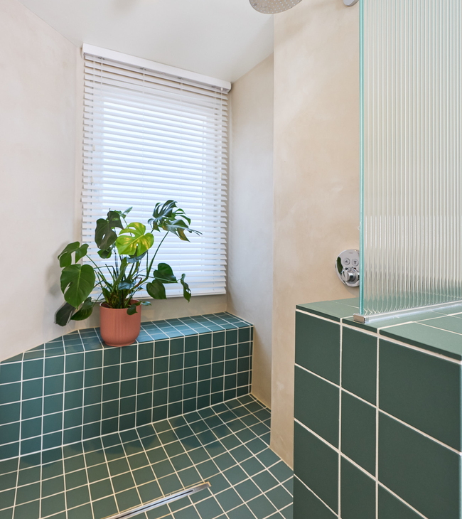 Douche met groene tegels en wit voegsel, ingebouwde zitbank, regendouche en plant bij raam met jaloezieën.
