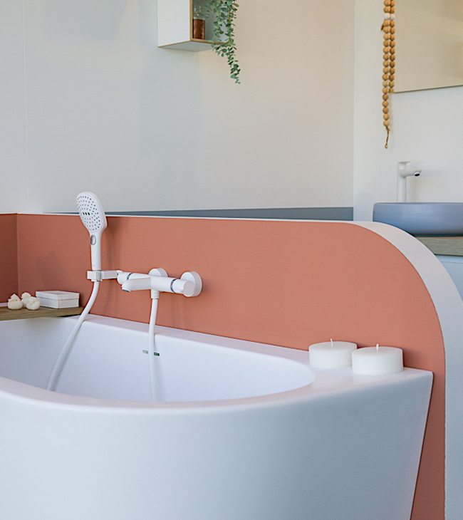 Vrijstaand hoekbad met afgeronde vorm, witte kranen en handdouche, omgeven door sfeervolle accessoires in een Nordic badkamer.